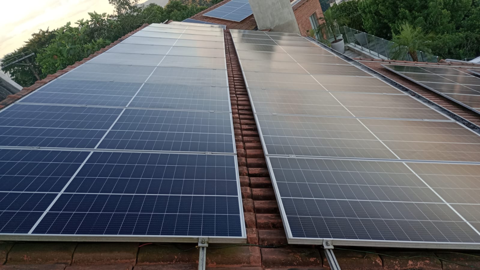 Fileiras de módulos fotovoltaicos instalados sobre uma cobertura, com vegetação ao fundo.